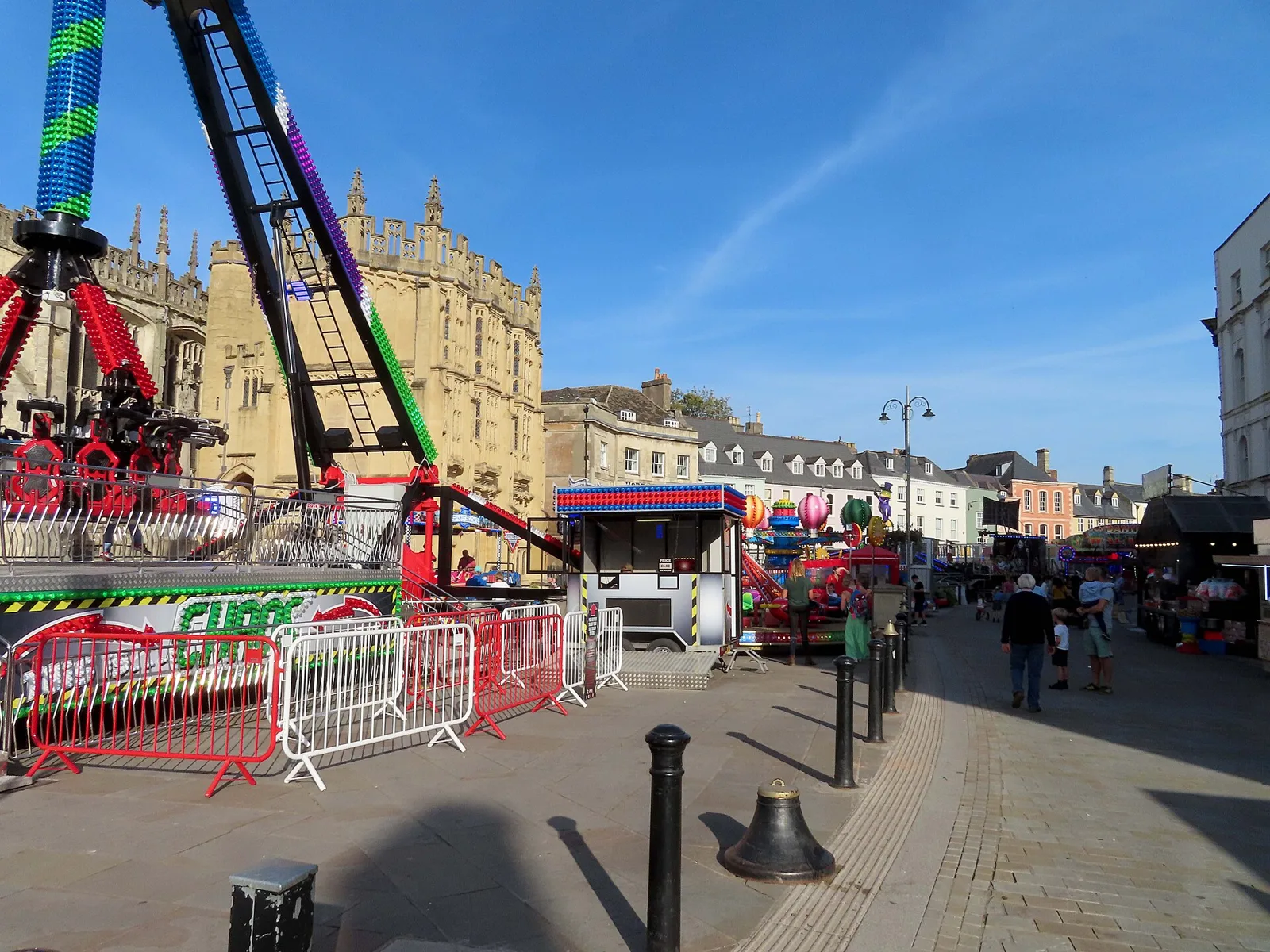 Fairground rides set up along a Cotswold town street beside a stone church, with people walking between them on a sunny evening